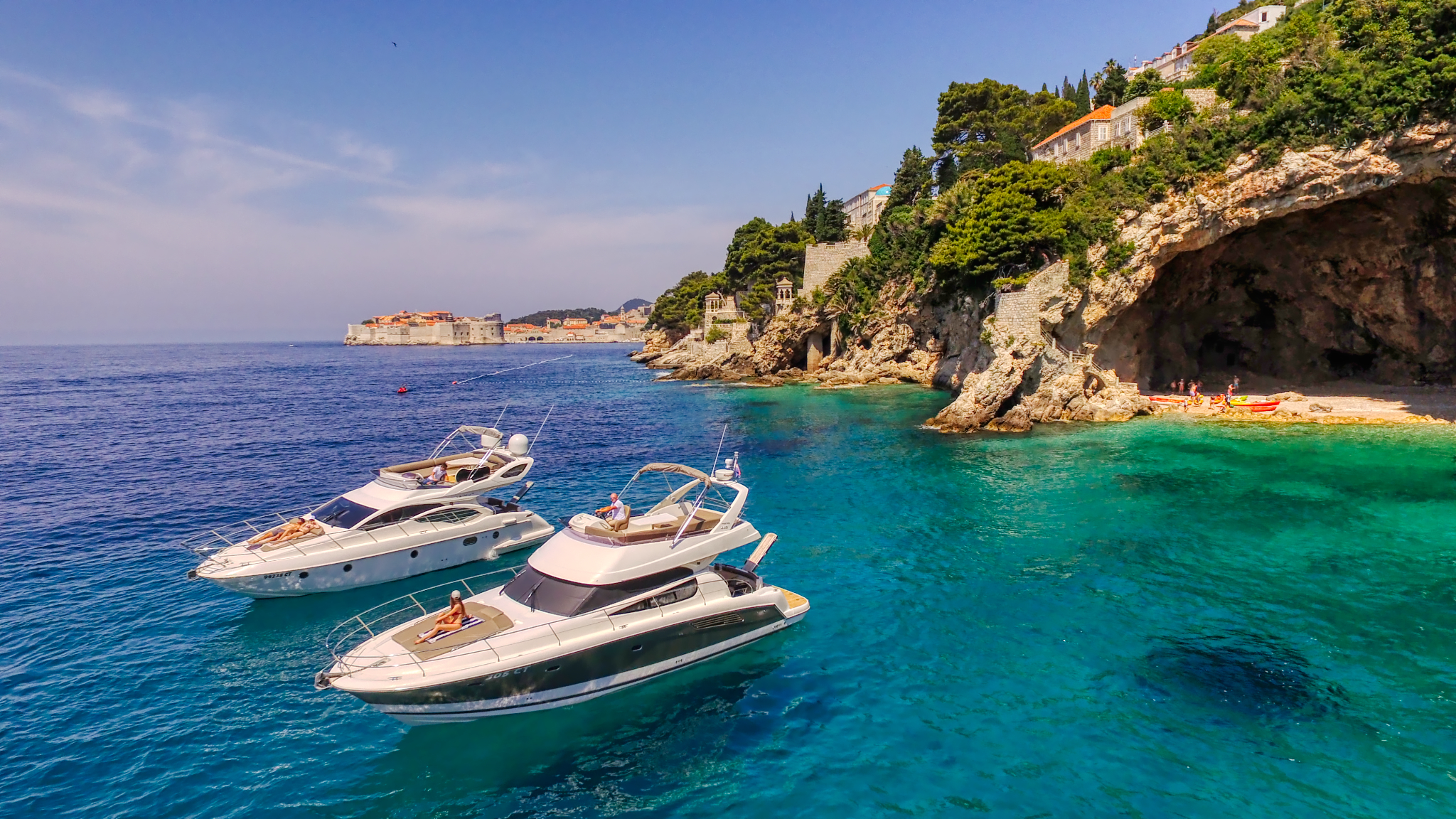 Two yachts of the Dubrovnik Boat Adventure fleet anchored in a crystal-blue cove with the Old Town walls in the distance
